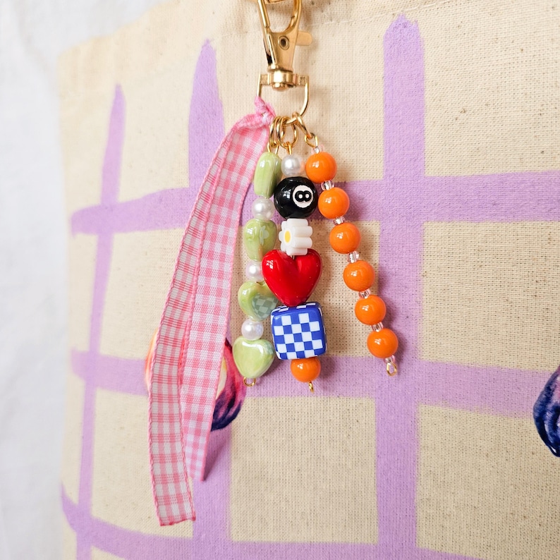 May include: A beaded charm with a gold-colored clasp and a pink and white gingham ribbon. The charm features a variety of beads, including green heart-shaped beads, a red heart, a blue and white checkered cube, and an eight-ball bead.