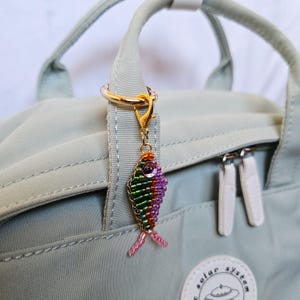 May include: A beaded fish charm in shades of green, purple, orange, and pink, attached to a gold-colored clasp. The charm is hanging from a light green backpack handle with a gold ring.