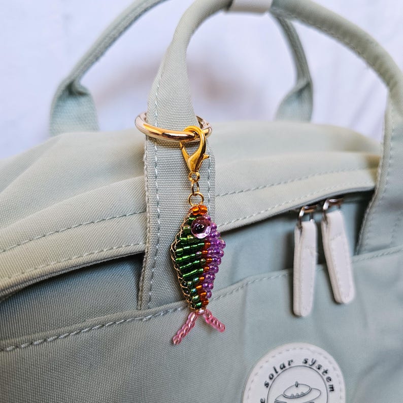 May include: A beaded fish charm in shades of green, purple, orange, and pink, attached to a gold-colored clasp. The charm is hanging from a light green backpack handle with a gold ring.