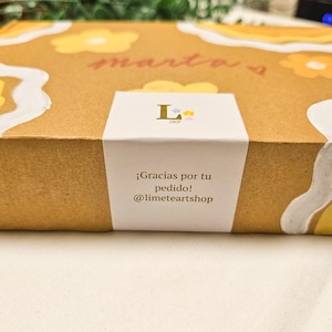 May include: A brown cardboard box with a white label. The box is decorated with yellow flower designs and the name "marta" in red script. The label reads "&iexcl;Gracias por tu pedido! @limeteartshop".