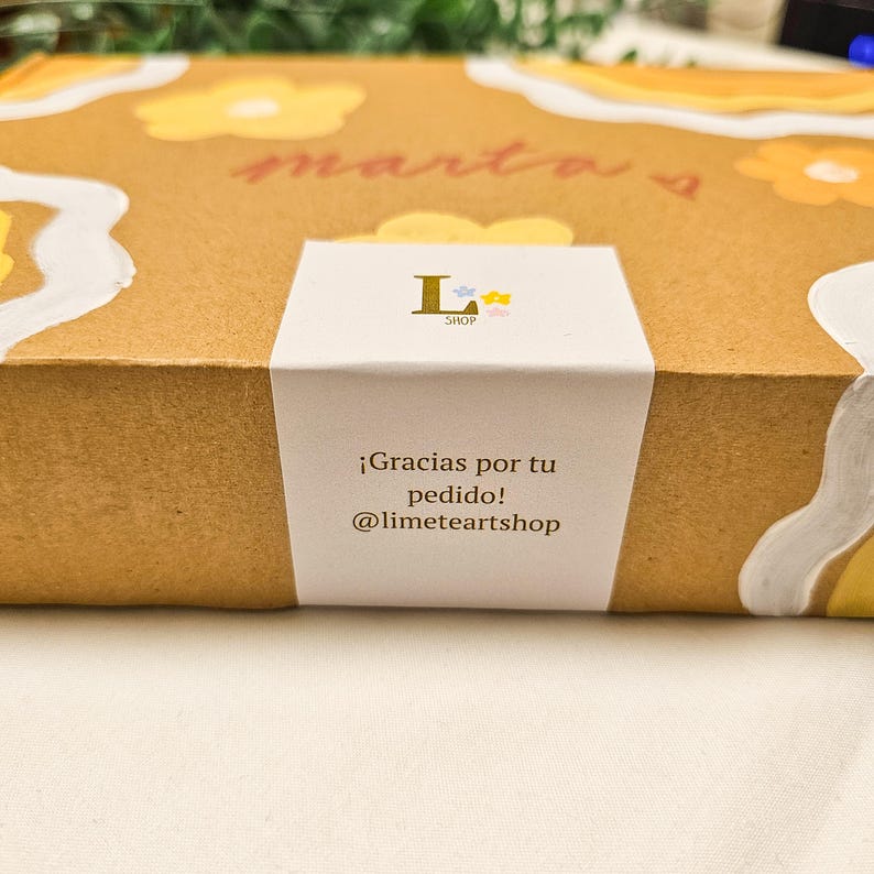 May include: A brown cardboard box with a white label. The box is decorated with yellow flower designs and the name "marta" in red script. The label reads "&iexcl;Gracias por tu pedido! @limeteartshop".