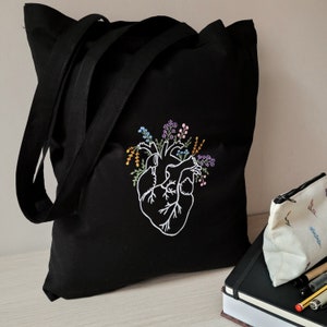 K&ouml;nnte beinhalten: Schwarze Einkaufstasche mit einem wei&szlig;en gestickten anatomischen Herzen, umgeben von bunten Blumen. Die Tasche hat zwei lange Henkel.