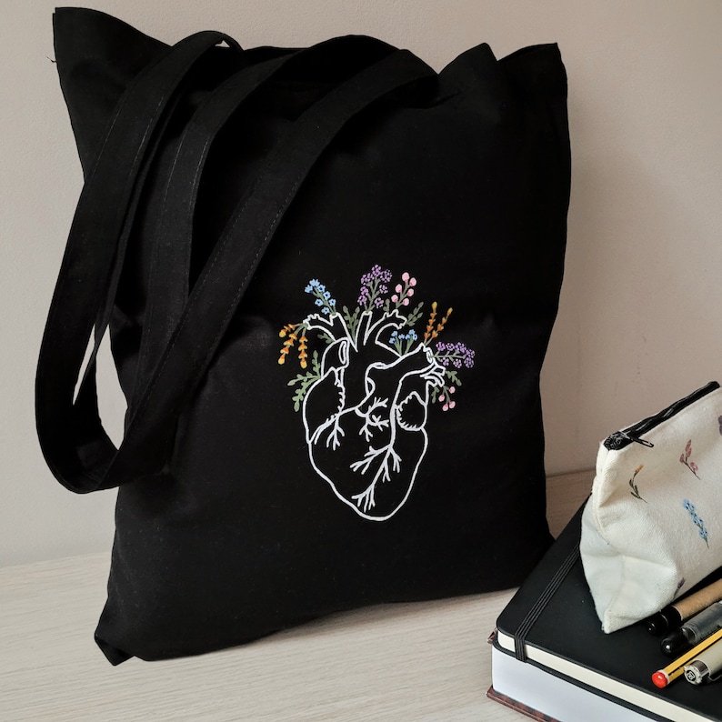 K&ouml;nnte beinhalten: Schwarze Einkaufstasche mit einem wei&szlig;en gestickten anatomischen Herzen, umgeben von bunten Blumen. Die Tasche hat zwei lange Henkel.