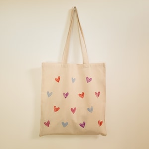 May include: A beige tote bag with a pattern of colorful hearts. The hearts are in shades of pink, purple, and blue.