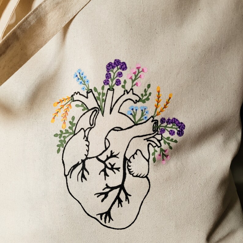 K&ouml;nnte beinhalten: Eine Tragetasche mit einem gestickten Design eines menschlichen Herzens, aus dem Blumen wachsen. Die Blumen sind in den Farben Lila, Rosa, Blau, Gelb und Gr&uuml;n.