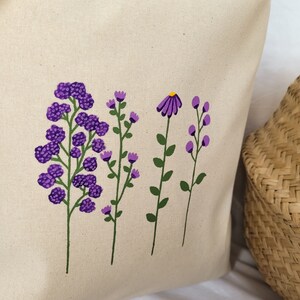 May include: A white tote bag with a purple floral design. The flowers are painted in a simple, stylized manner. The bag is made of canvas and has a natural, unbleached look.