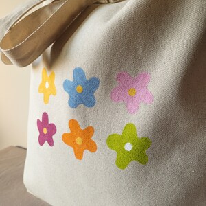 Peut inclure: Un sac fourre-tout en toile beige avec six motifs de fleurs color&eacute;s. Les fleurs sont dans des tons de rose, bleu, orange, jaune et vert.