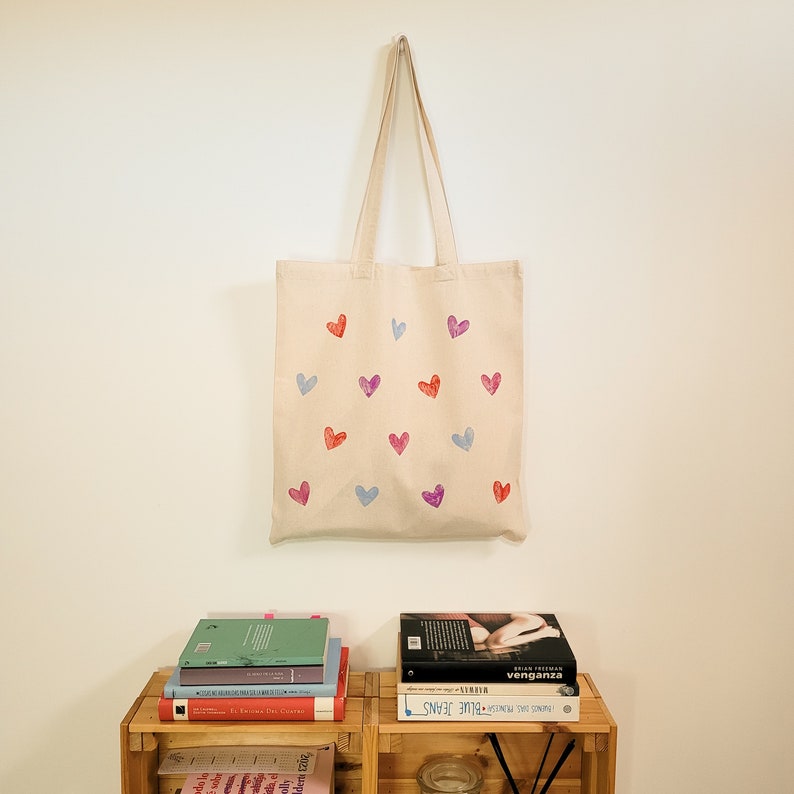 May include: A beige tote bag with a design of pink, purple, and red hearts. The bag is hanging on a white wall.