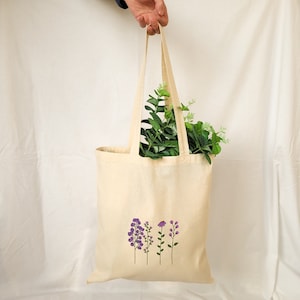 May include: A beige tote bag with a design of purple flowers on the front. The bag has a long handle and is made of canvas.