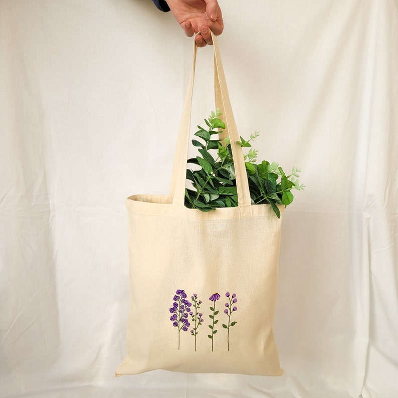 May include: A beige tote bag with a design of purple flowers on the front. The bag has a long handle and is made of canvas.