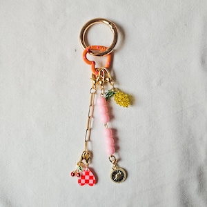 May include: A gold key chain with a pink and orange heart charm, pink beads, a pineapple charm, a checkered heart charm, and a gold circle charm.