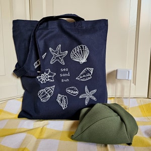 May include: A dark blue tote bag with white illustrations of seashells, starfish, and the words "sea sand sun". The bag is on a yellow and white checked surface with a green hat and a clear plastic bottle.