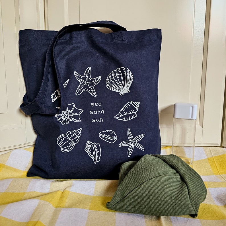 May include: A dark blue tote bag with white illustrations of seashells, starfish, and the words "sea sand sun". The bag is on a yellow and white checked surface with a green hat and a clear plastic bottle.