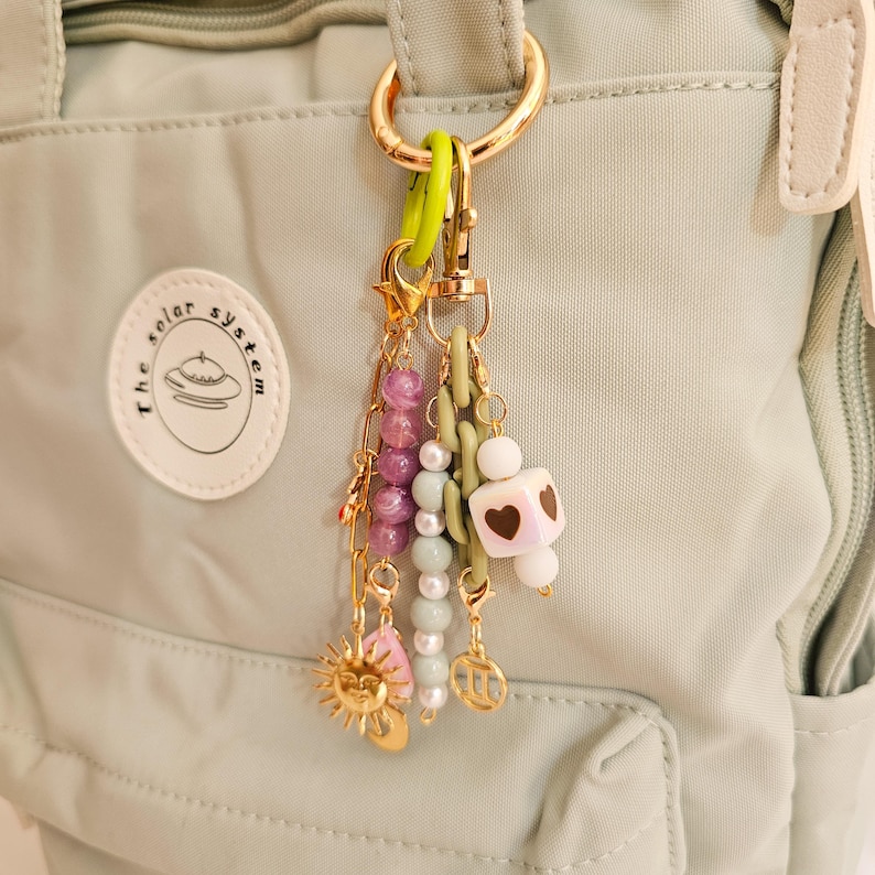May include: A gold-toned keychain with various charms, including a sun, a pink leaf, and a white cube with heart cutouts. The keychain also features strands of purple and white beads, and a green rubber ring.