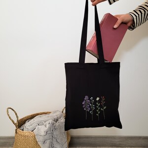 K&ouml;nnte beinhalten: Schwarze Tragetasche mit einem floralen Stickmuster. Die Tasche ist aus Canvas und hat einen langen Henkel. Ein Buch liegt auf der Tasche.