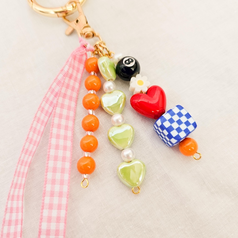 May include: A beaded keychain with a gold clasp and pink gingham ribbon. The charm features orange beads, green heart-shaped beads, a red heart, a blue and white checkered cube, and a black eight-ball.