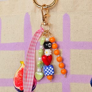 May include: A gold-toned keychain with a charm featuring colorful beads. The charm includes a black 8-ball bead, a red heart, a checkered cube, and orange beads. A pink and white gingham ribbon is attached.