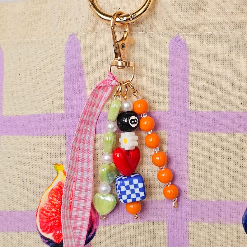 May include: A gold-toned keychain with a charm featuring colorful beads. The charm includes a black 8-ball bead, a red heart, a checkered cube, and orange beads. A pink and white gingham ribbon is attached.