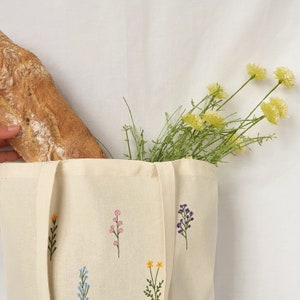 May include: A white canvas tote bag with embroidered floral designs. The bag is filled with a loaf of bread and a bouquet of yellow flowers.