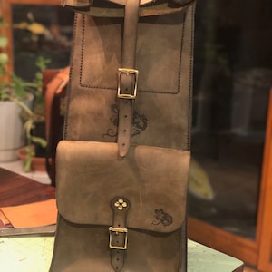 Könnte beinhalten: Eine braune Ledertasche mit zwei Schnallen und einer Klappe. Die Tasche hat ein Blumenmuster und das Wort "Profesa" steht unten.