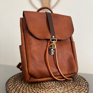 Op de afbeelding: Een bruine leren rugzak met een donkerbruine leren band en een gouden metalen sluiting. De rugzak heeft een klein vakje aan de voorkant en de tekst "PROFESSOR - LEATHER -" is gedrukt op de onderste band.