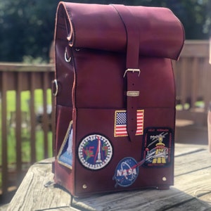 May include: A burgundy leather backpack with a roll-top closure and multiple patches, including a US flag, NASA logo, and astronaut patches.
