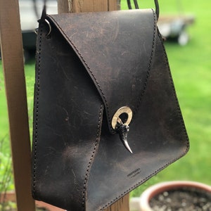 May include: A dark brown leather crossbody bag with a silver buckle and a silver horn-shaped accent. The bag has a flap closure and a strap that can be adjusted. The bag is made by Professora Leather.