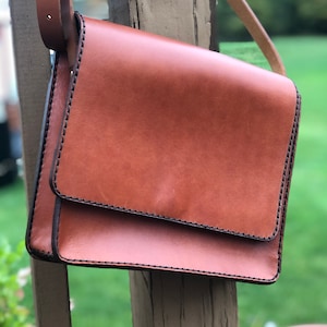 May include: A brown leather crossbody bag with a flap closure. The bag has black stitching around the edges and a long adjustable strap. The bag is hanging on a wooden post with a blurred green background.