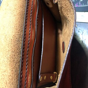 May include: Close-up of a handcrafted brown leather bag, revealing a zipped pocket and internal compartments. The interior is lined with a lighter brown suede. Brass hardware and detailed stitching are visible.
