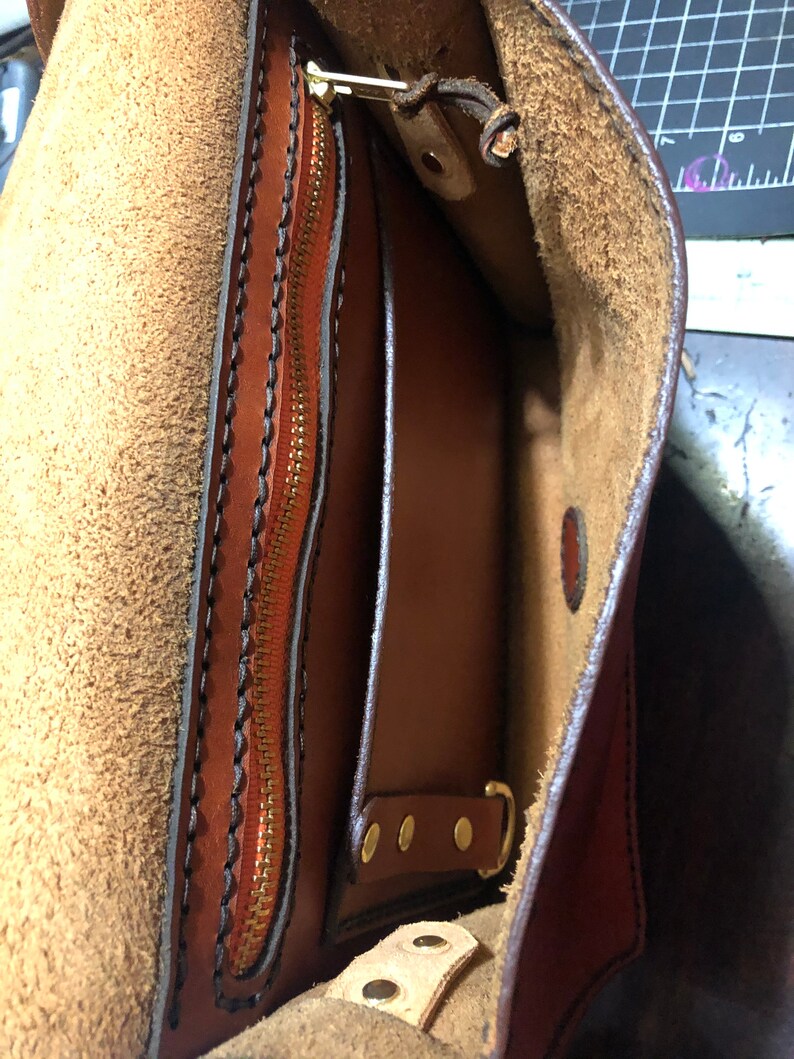 May include: Close-up of a handcrafted brown leather bag, revealing a zipped pocket and internal compartments. The interior is lined with a lighter brown suede. Brass hardware and detailed stitching are visible.