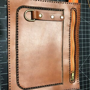 May include: A handcrafted brown leather pouch with a stitched border and a brass-coloured metal D-ring. It features a vertical orange zip with a brass-coloured pull and a small leather strap with rivets.