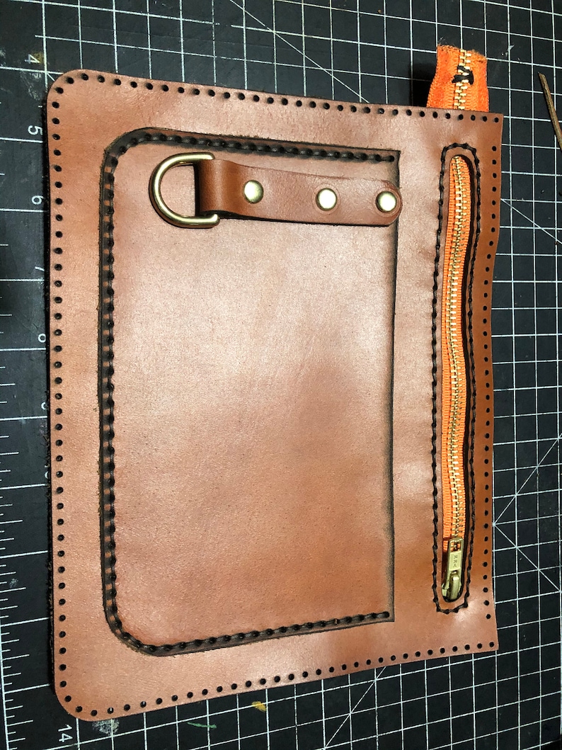 May include: A handcrafted brown leather pouch with a stitched border and a brass-coloured metal D-ring. It features a vertical orange zip with a brass-coloured pull and a small leather strap with rivets.