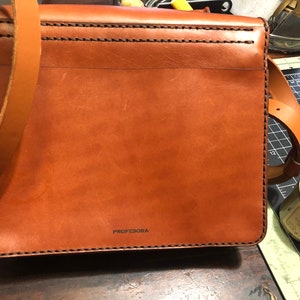 May include: A brown leather messenger bag with a long shoulder strap. The bag has visible stitching and the word "PROFESORA" printed on the front. The bag is rectangular and appears to be new.