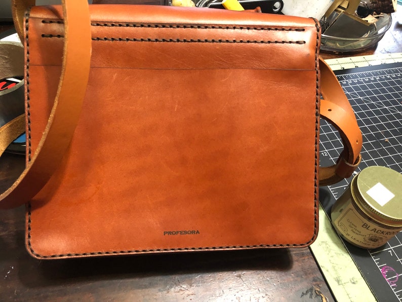 May include: A brown leather messenger bag with a long shoulder strap. The bag has visible stitching and the word "PROFESORA" printed on the front. The bag is rectangular and appears to be new.