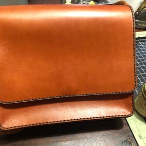 May include: A brown leather satchel with a flap closure and shoulder strap. The bag has black stitching along the edges and a smooth finish. The rectangular bag is likely new. The bag's dimensions are approximately 25 cm x 20 cm.