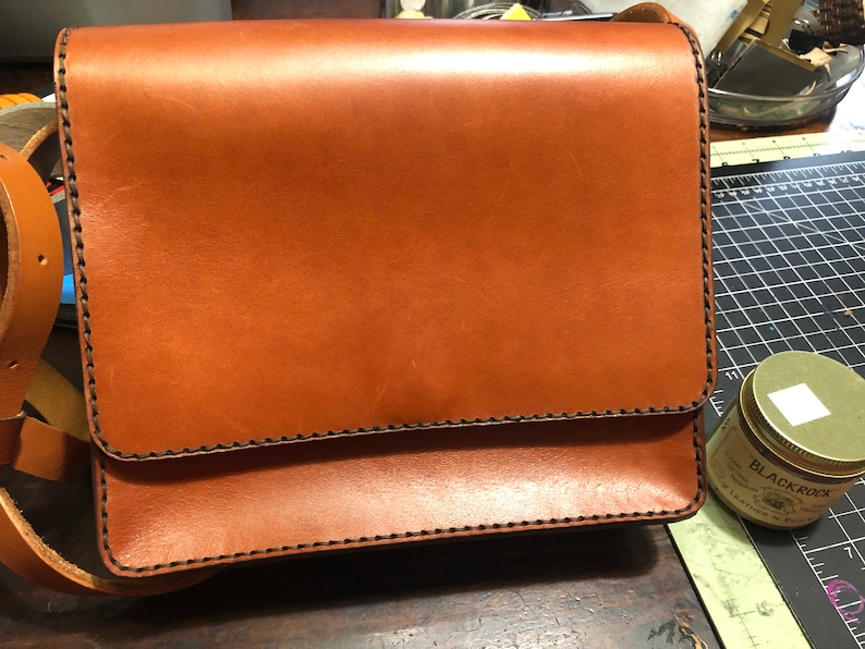 May include: A brown leather satchel with a flap closure and shoulder strap. The bag has black stitching along the edges and a smooth finish. The rectangular bag is likely new. The bag's dimensions are approximately 25 cm x 20 cm.