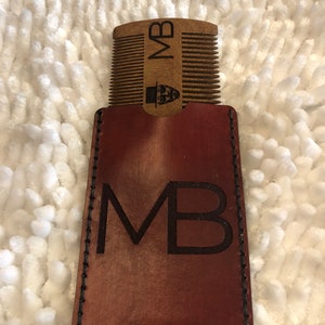 May include: A brown leather case with the initials "MB" embossed on the front. The case has a stitched border and holds a wooden comb with the same initials and a small beard icon.