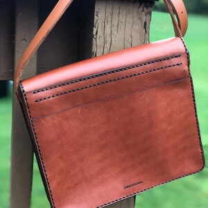 May include: A brown leather crossbody bag with a long strap. The bag is rectangular with a flap closure and black stitching. The word "PROFESORA" is printed on the lower right corner of the bag.