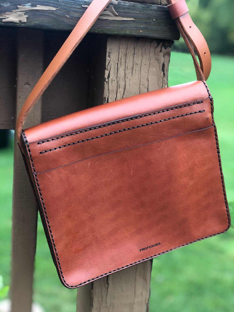 May include: A brown leather crossbody bag with a long strap. The bag is rectangular with a flap closure and black stitching. The word "PROFESORA" is printed on the lower right corner of the bag.