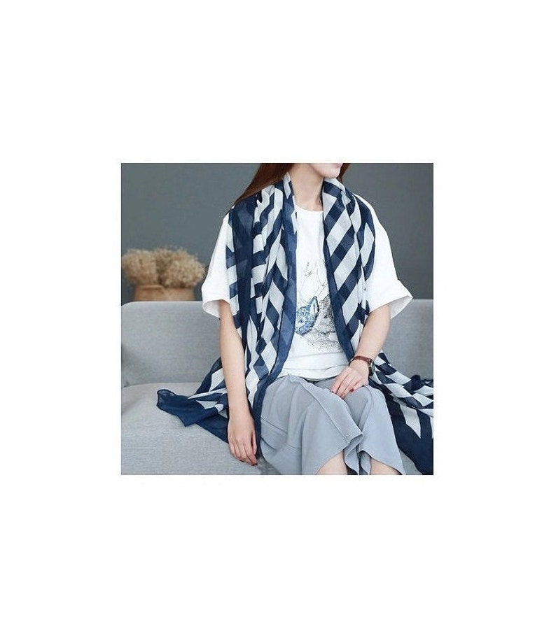 Puede incluir: Una bufanda a rayas azul marino y blanca sobre una camiseta blanca y pantalones grises. La bufanda tiene un patr&oacute;n geom&eacute;trico audaz. La persona est&aacute; sentada en un sof&aacute; gris.