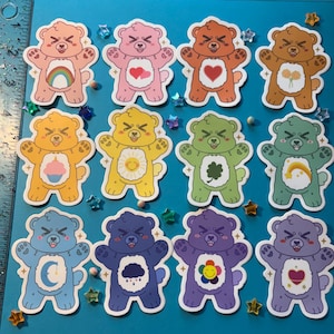 May include: A collection of twelve colorful bear stickers with various designs. Each bear has a unique color and a symbol on its belly, such as a rainbow, heart, or flower. The stickers are arranged on a blue surface with star-shaped decorations.
