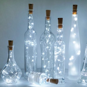 May include: A set of clear glass bottles with cork tops filled with white fairy lights. The bottles are various shapes and sizes, and the lights are strung throughout the bottles.