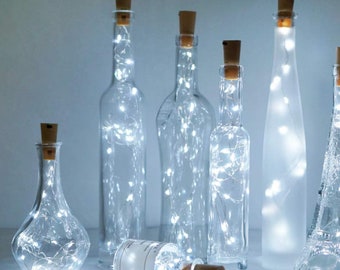 May include: A set of clear glass bottles with cork tops filled with white fairy lights. The bottles are various shapes and sizes, and the lights are strung throughout the bottles.