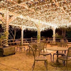 May include: A patio with a wooden pergola covered in white string lights. The patio is furnished with wicker chairs and tables.