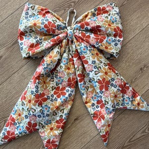 May include: A large fabric hair bow featuring a repeating floral pattern with red, yellow, and blue flowers on a white background. The bow has two wide, pointed tails extending downward and a central knot with a small fabric loop at the top.