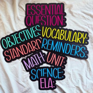 May include: A collection of colorful classroom decor signs with black backgrounds and various text. The signs include phrases like "Essential Question," "Objectives," "Vocabulary," and "Math Unit." The text is in bright colors like pink, yellow, and blue.