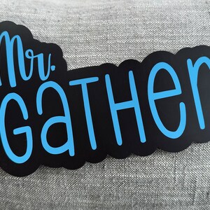 Puede incluir: Pegatina de vinilo negro y azul con el texto "Mr. Gathen" en una fuente cursiva.