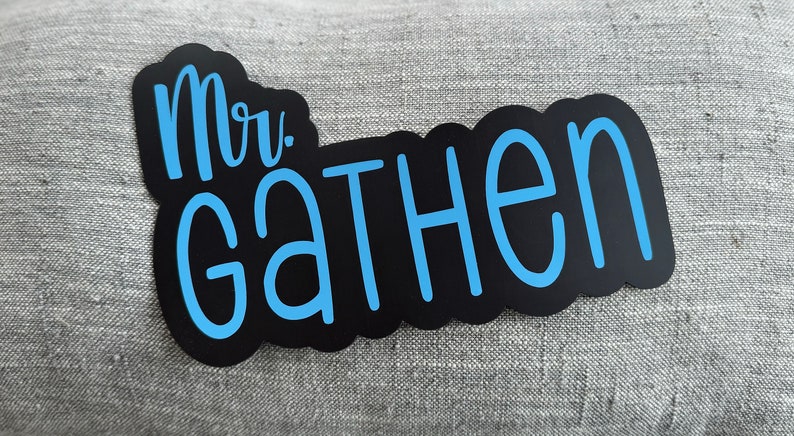 Puede incluir: Pegatina de vinilo negro y azul con el texto "Mr. Gathen" en una fuente cursiva.
