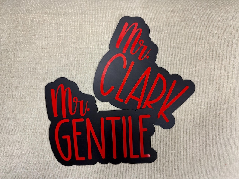 Puede incluir: Dos letreros negros con letras rojas. Los letreros dicen "Mr. Clark" y "Mr. Gentile".