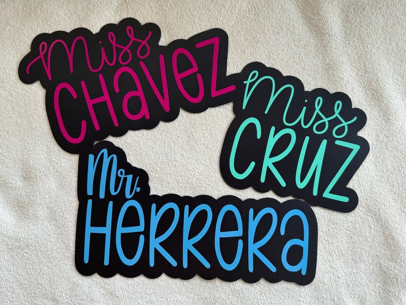 Puede incluir: Tres etiquetas de nombre negras con letras blancas. Las etiquetas de nombre dicen "Miss Chavez", "Miss Cruz" y "Mr. Herrera". Las letras est&aacute;n en una fuente cursiva. Las etiquetas de nombre son de diferentes colores: rosa, turquesa y azul.